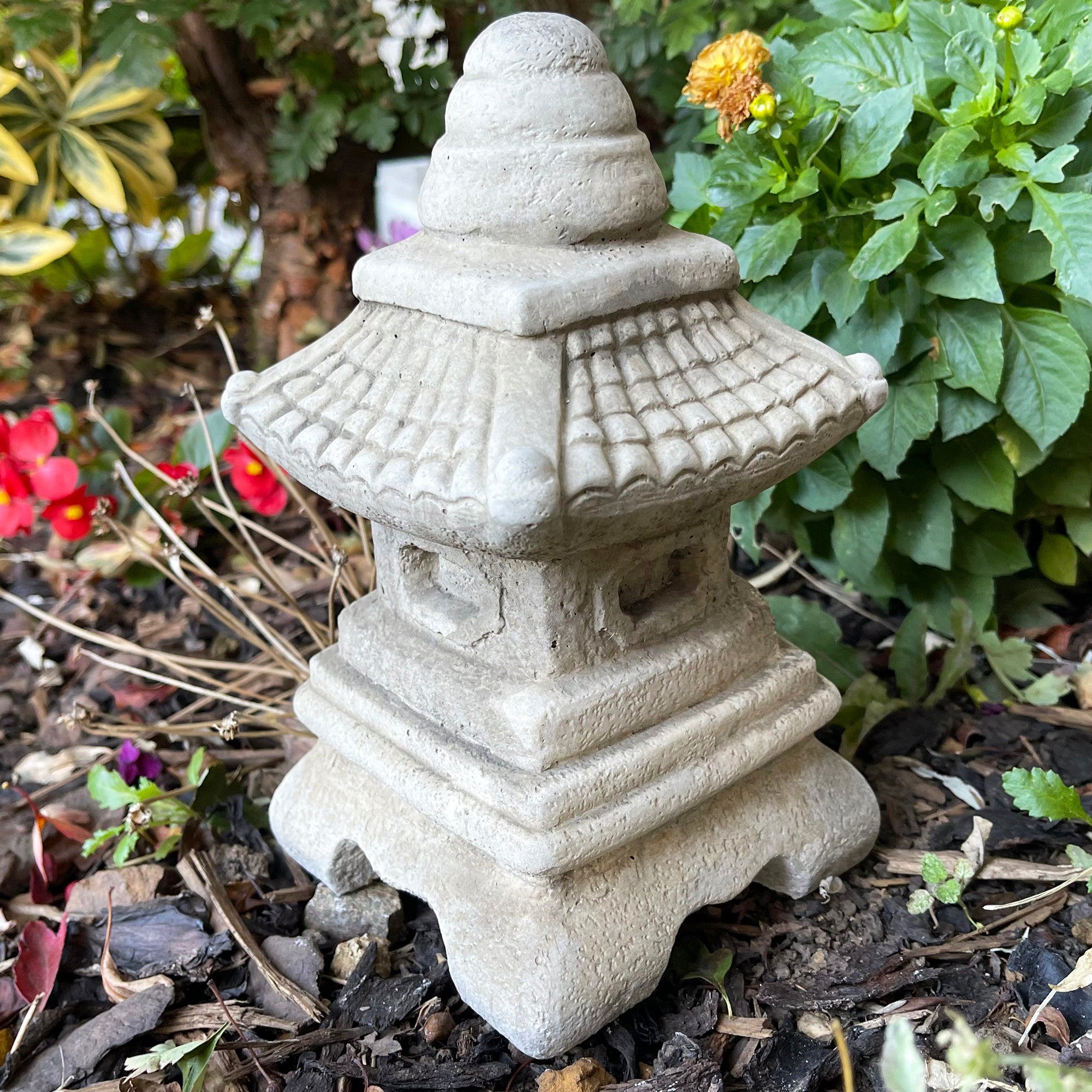 A traditional Japanese pagoda garden sculpture made of stone with an antique grey finish, situated in a garden setting with plants around it.