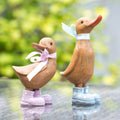 Two wooden duck figurines with bows and boots on a reflective surface with a blurred natural background.