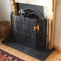 A black cast iron fire screen with Fleur-de-Lis design is positioned in front of a fireplace, with the fireplace visible in the background.