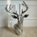A silver-colored wall-mounted sculpture of a stags head with large antlers, placed against an indoor background.