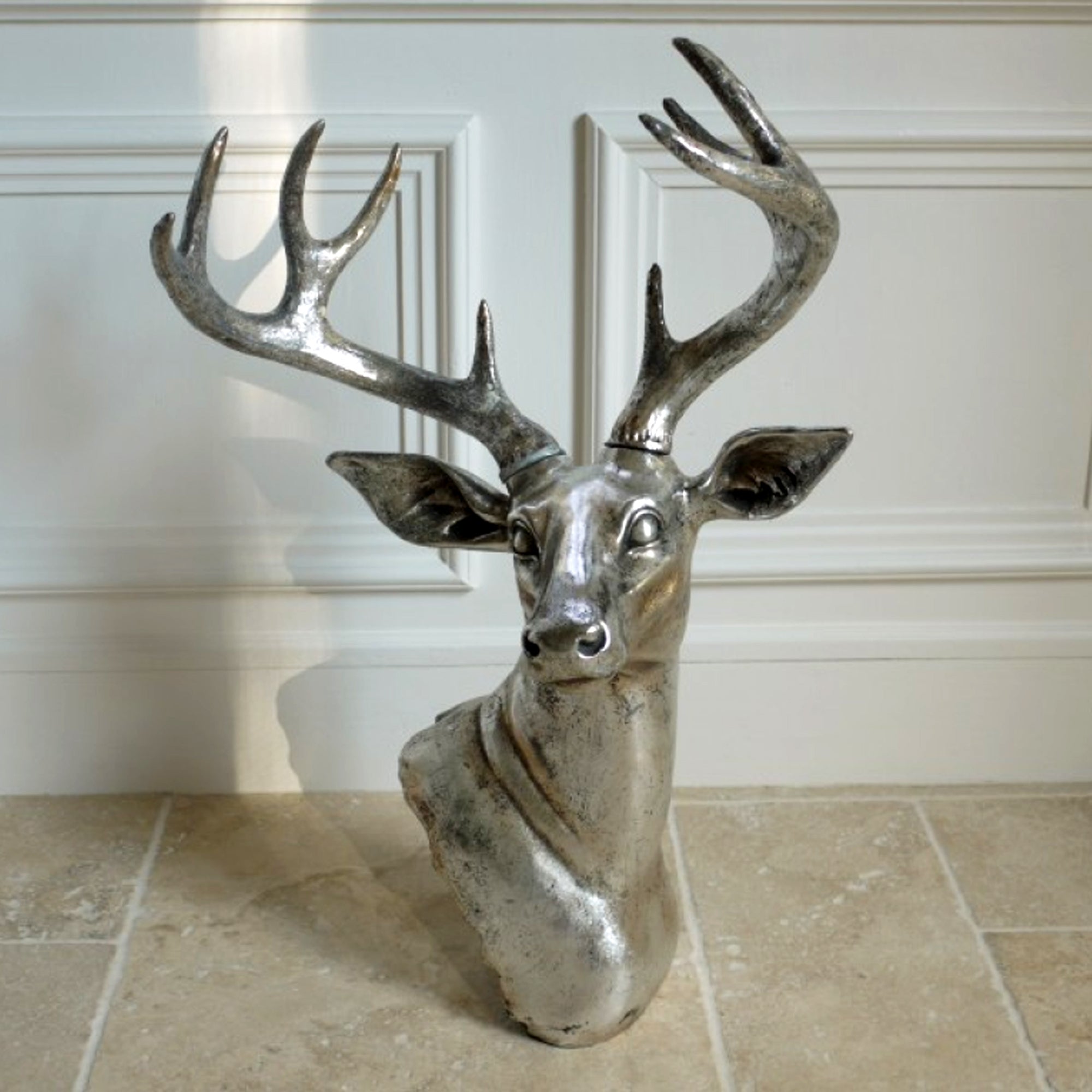 A silver-colored wall-mounted sculpture of a stags head with large antlers, placed against an indoor background.