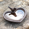 A cast iron heart-shaped bird bath with a screwable robin bird perched on the edge, situated on a wooden surface.