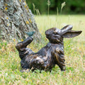 Bronze sculpture of a rabbit with a bird on its back in a grassy outdoor setting.