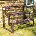 A brown cast iron rectangle plant stand with a 3-tier staircase design, featuring intricate swirl scrollwork, placed on grass against a stone wall.
