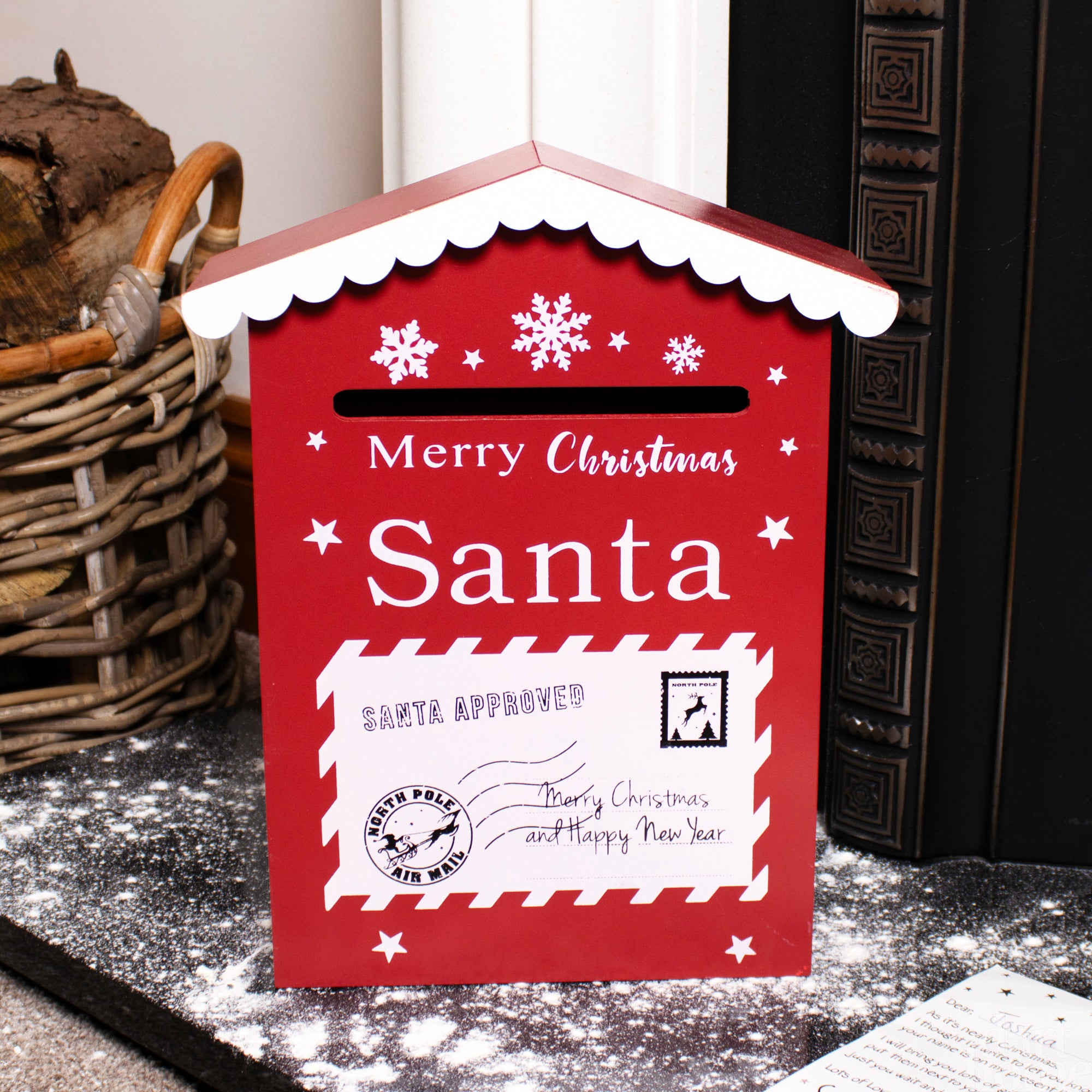 A house-shaped red and white Christmas themed post box with 'Merry Christmas Santa' text and a white Santa Approved letter design, featuring star and snowflake motifs, placed on a table with a wreath in the background.