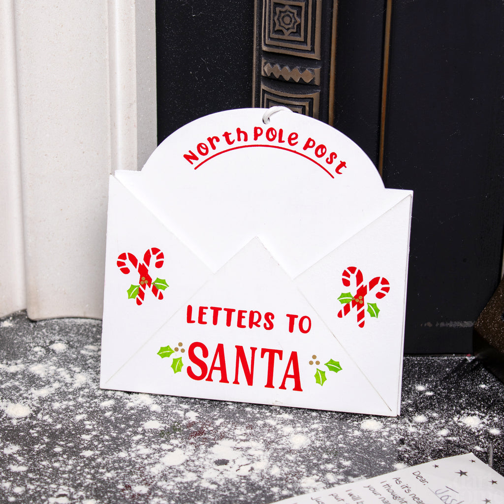 A white envelope-shaped Christmas decor piece with a 'NORTH POLE POST TO SANTA' design, featuring red candy cane and hollyberry details, placed against a fireplace backdrop.