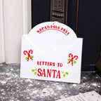 A white envelope-shaped Christmas decor piece with a 'NORTH POLE POST TO SANTA' design, featuring red candy cane and hollyberry details, placed against a fireplace backdrop.