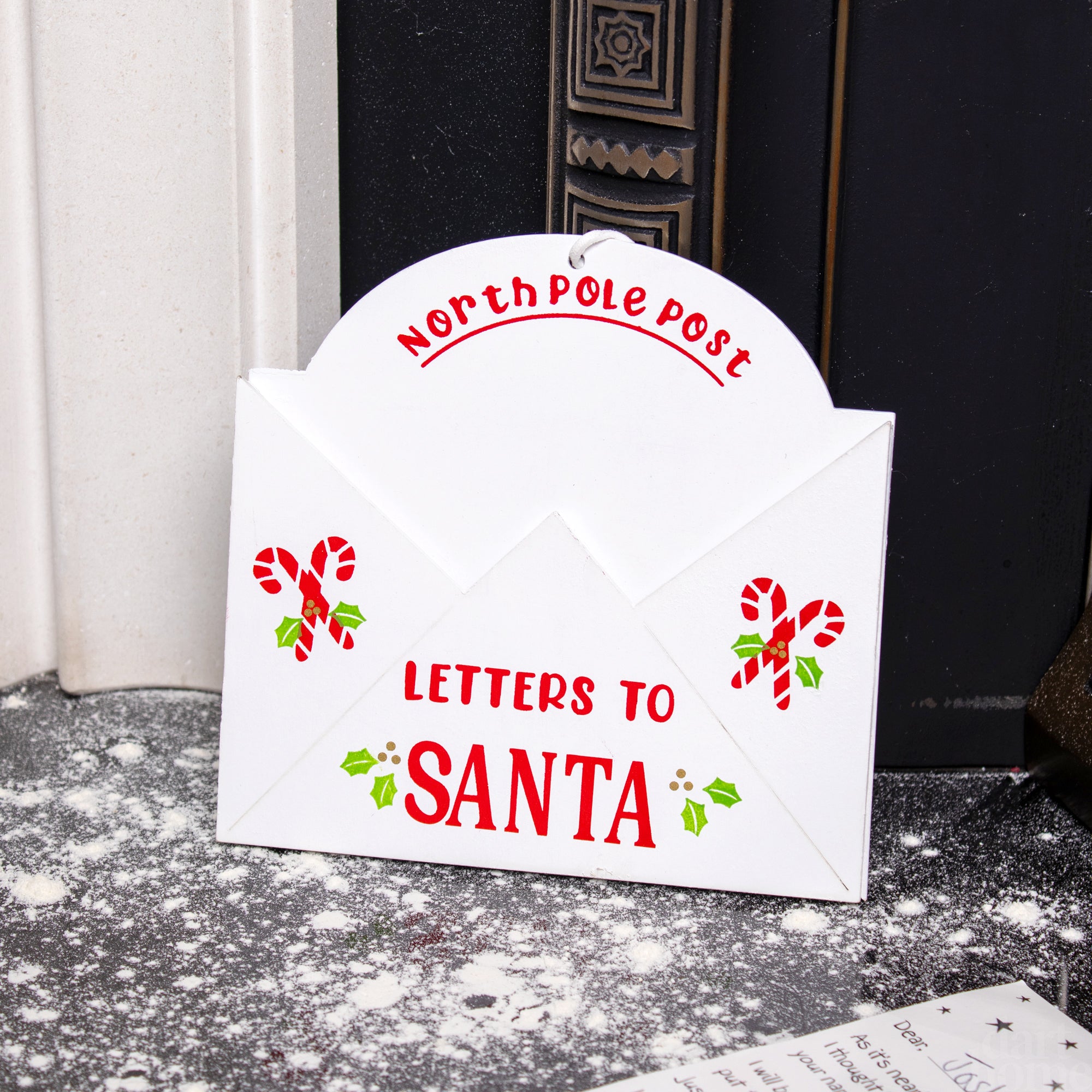 A white envelope-shaped Christmas decor piece with a 'NORTH POLE POST TO SANTA' design, featuring red candy cane and hollyberry details, placed against a fireplace backdrop.