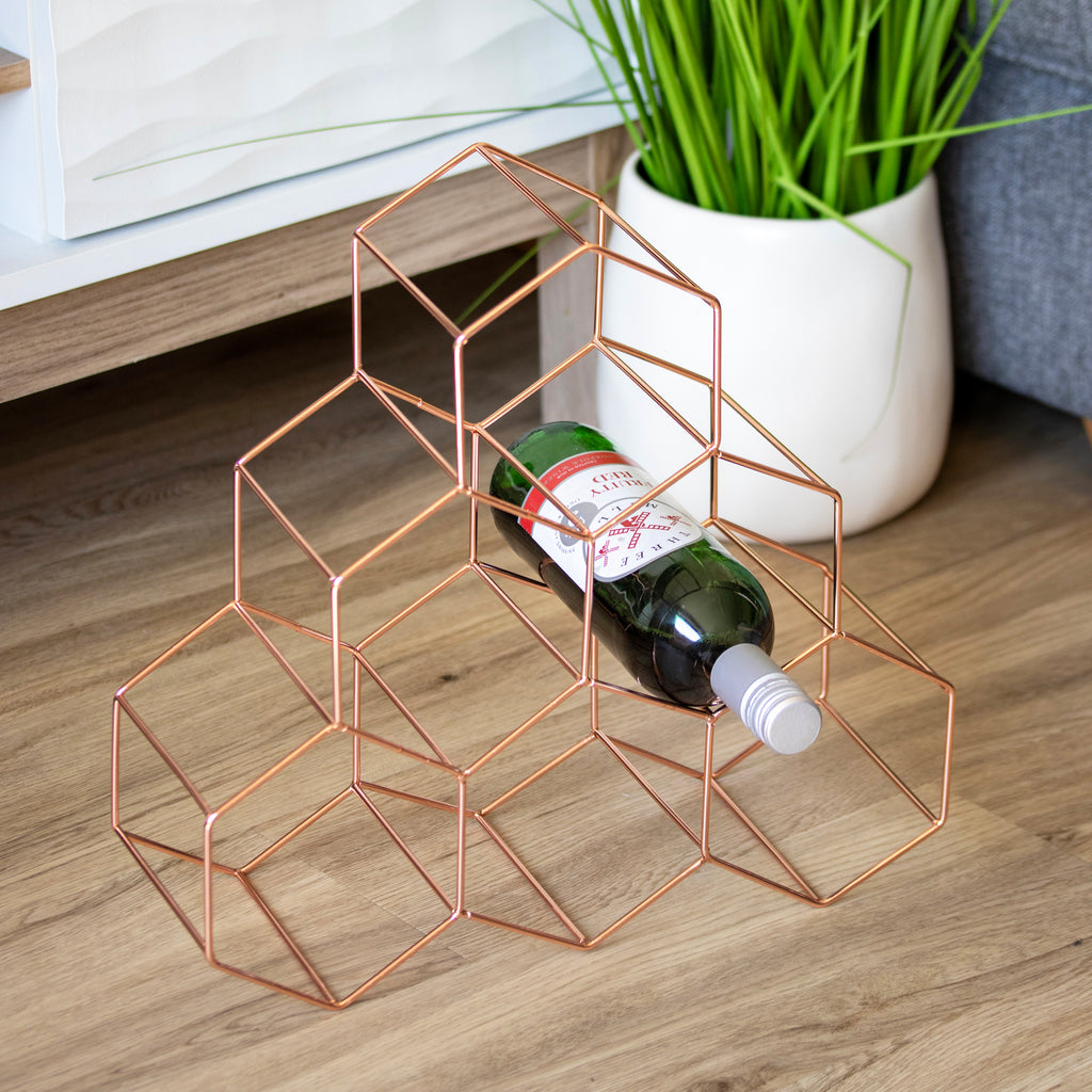 Geometric copper wine rack holding a bottle on a wooden surface with a plant in the background.