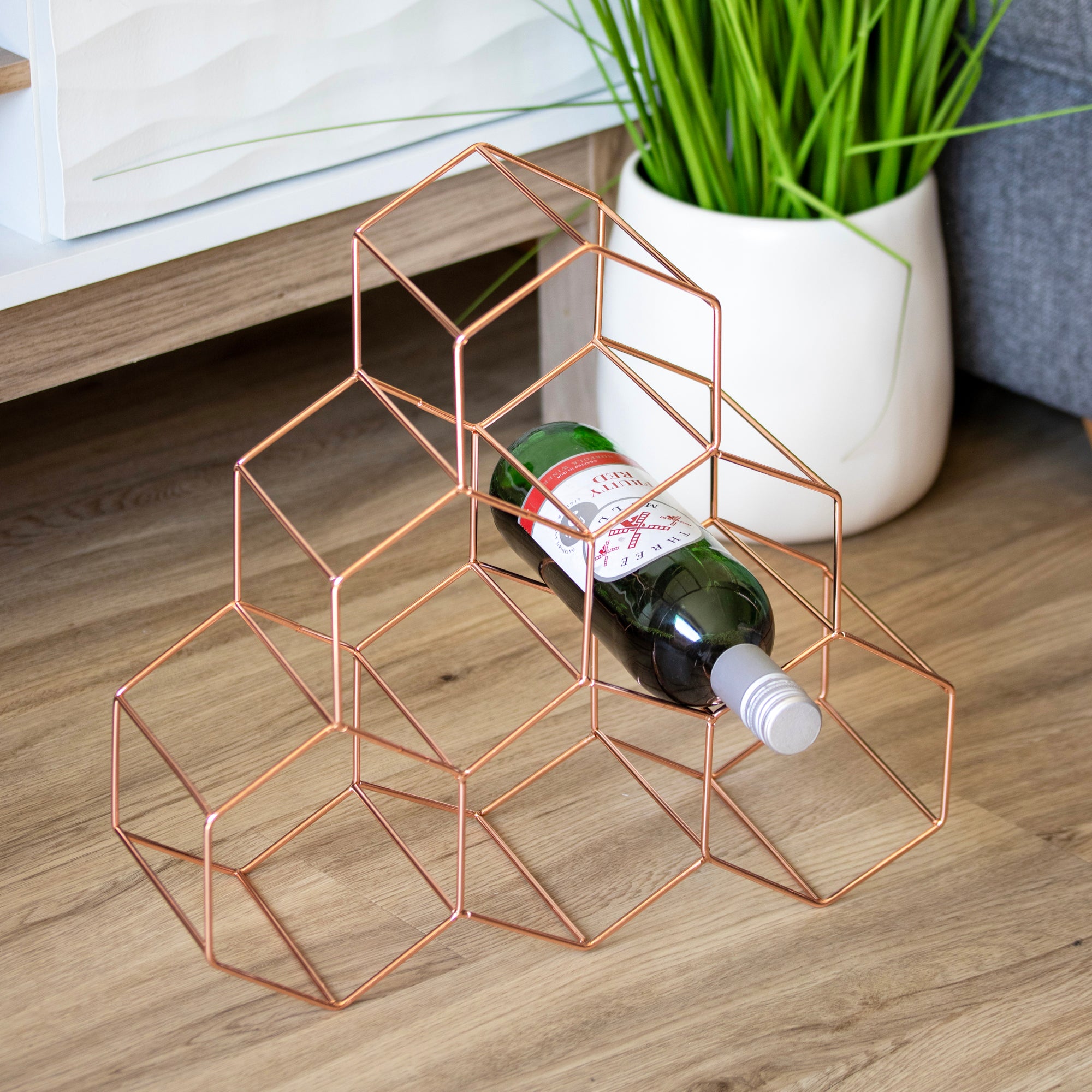 Geometric copper wine rack holding a bottle on a wooden surface with a plant in the background.