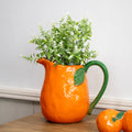 Orange pitcher with green handle and leaves holding plants on a wooden surface.