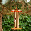 Copper bird seed feeder with 6 feeding ports, hanging from a tree branch with a blurred green foliage background