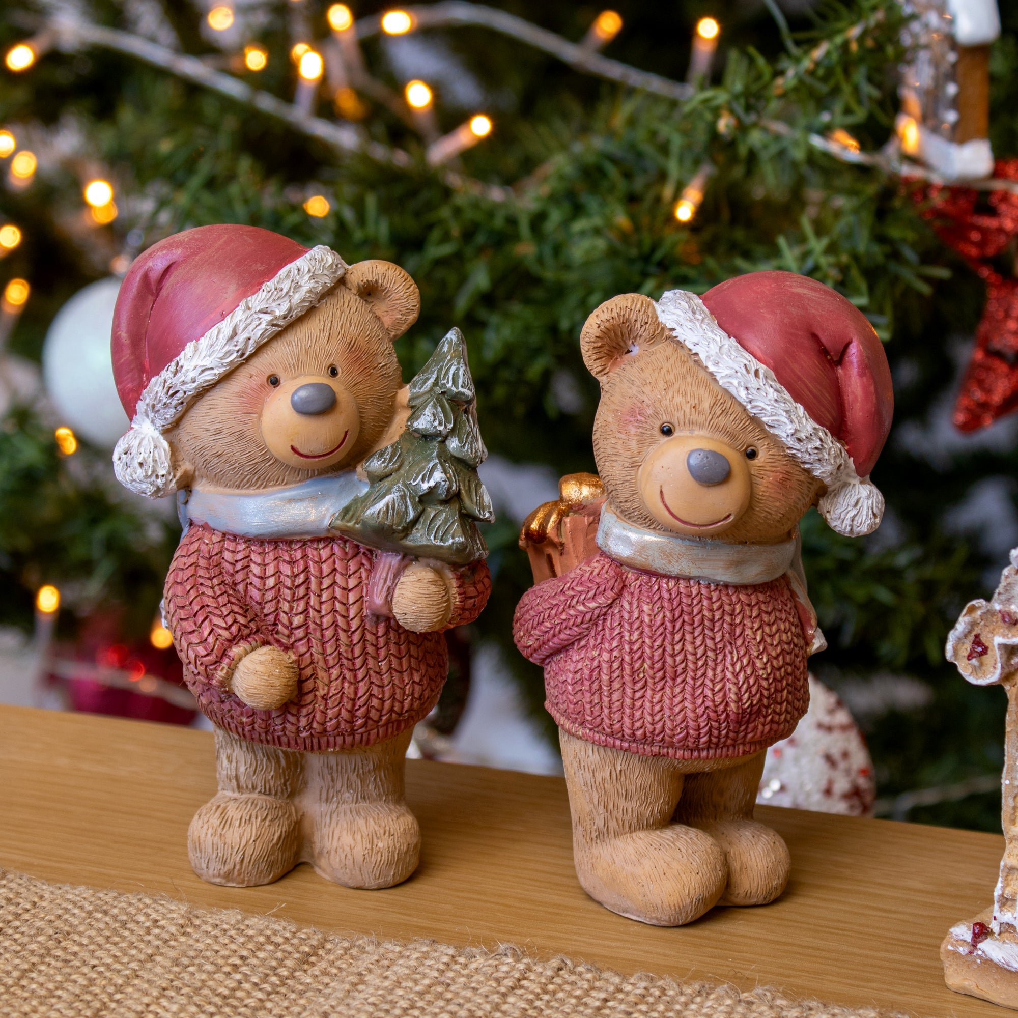 Two teddy bear figurines wearing Christmas hats and holding decorative branches, placed on a table with a blurred Christmas tree and gifts in the background.