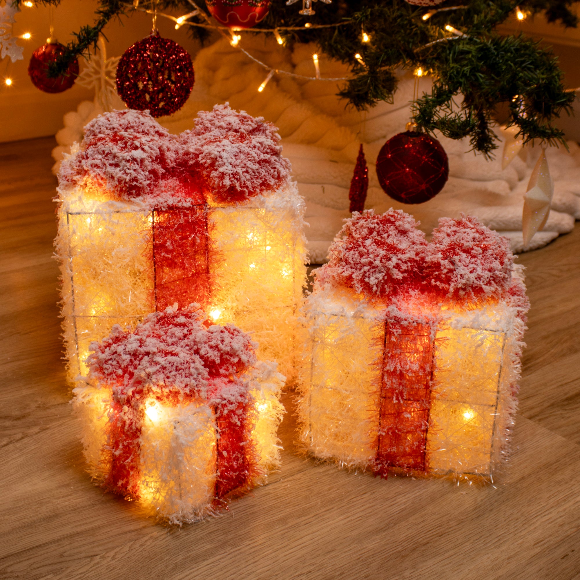 Set of three LED light-up Christmas present decorations with a white and red design, artificial snow, and red bow details, displayed on a wooden floor with a Christmas tree in the background.
