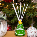 A hand-painted, Christmas tree-shaped ceramic diffuser bottle with multiple reed sticks inserted on top, decorated with a yellow star, set against a festive holiday backdrop.