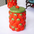 A red ceramic kitchen storage jar with a textured strawberry fruit design and a green lid, displayed on a white surface.