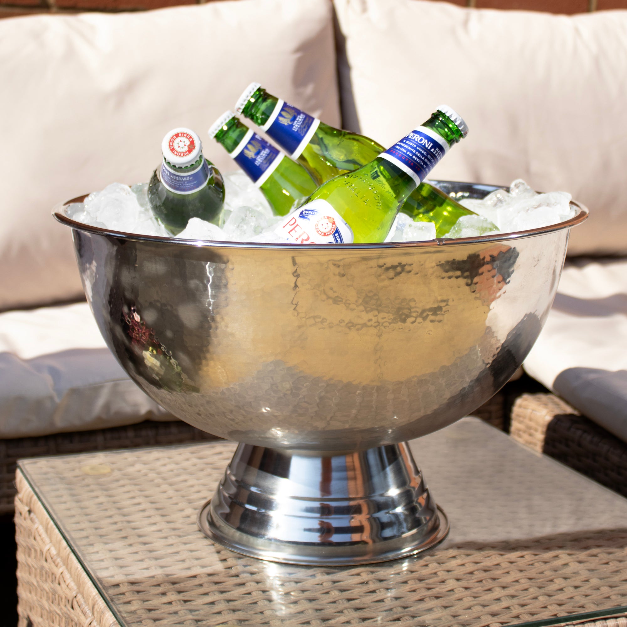A large hammered stainless steel champagne bucket filled with ice and bottles, placed on a glass table with a wicker sofa in the background.