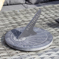 A round gray slate sundial with Roman numeral detailing and the word 'SUNDIAL' engraved on it, placed on a wicker surface.