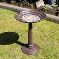 An extra large brown cast iron pillar bird bath with a round bowl mounted on a pedestal base, featuring a perched bird detail, situated on a grass lawn with flowers in the background.