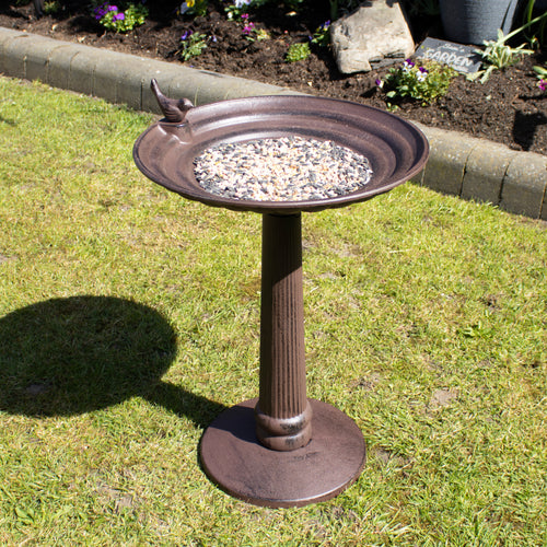 An extra large brown cast iron pillar bird bath with a round bowl mounted on a pedestal base, featuring a perched bird detail, situated on a grass lawn with flowers in the background.