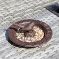 A brown cast iron sundial bird bath with a raised Roman numeral clock face and a perched bird detail, filled with bird seed, placed on a wicker surface.