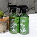 Two green glass spray bottles with white wildflower prints, fitted with black plastic triggers, displayed on a table with a plant in the background.