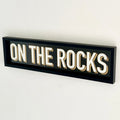 A black wood framed wall mounted 'On The Rocks' bar sign with block typography and a 3D effect, featuring white and gold colours.