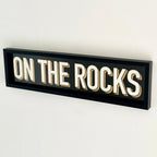 A black wood framed wall mounted 'On The Rocks' bar sign with block typography and a 3D effect, featuring white and gold colours.