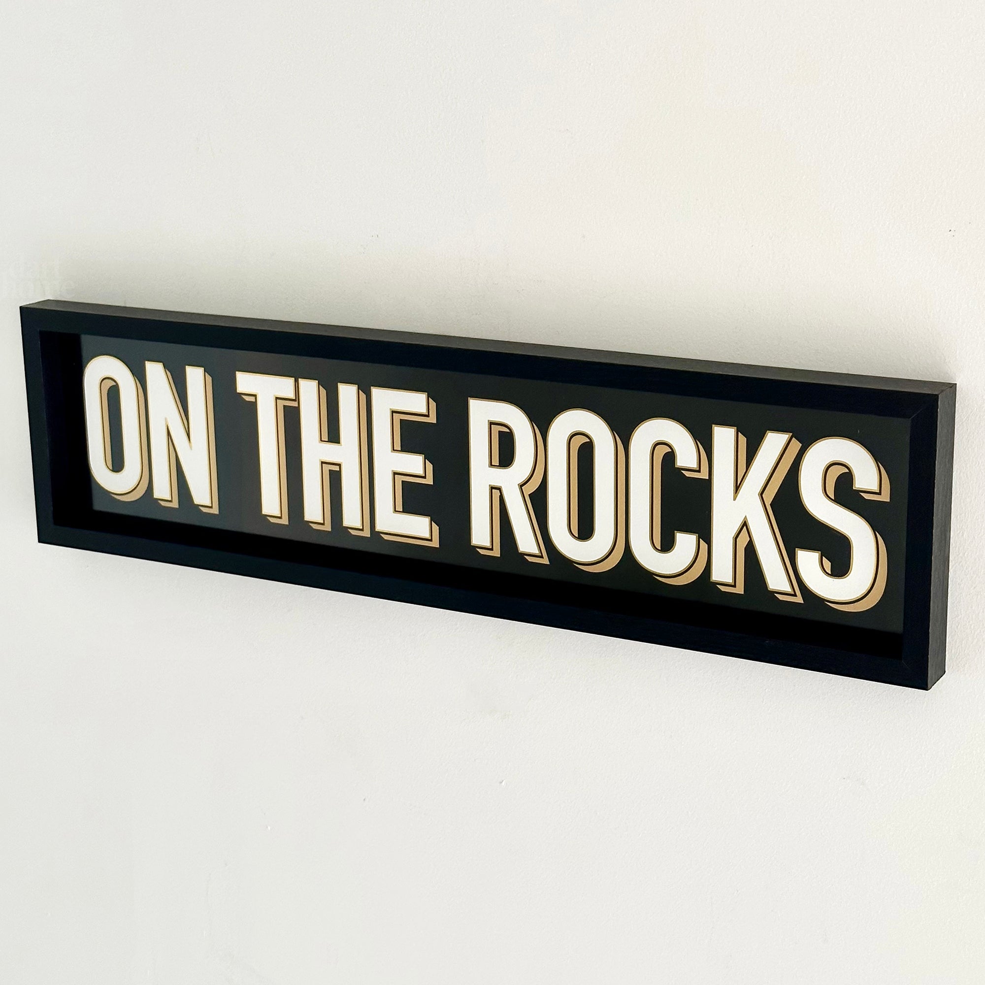 A black wood framed wall mounted 'On The Rocks' bar sign with block typography and a 3D effect, featuring white and gold colours.