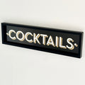 A black framed wall mounted 'COCKTAILS' sign with gold block typography against a white background.