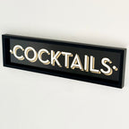 A black framed wall mounted 'COCKTAILS' sign with gold block typography against a white background.