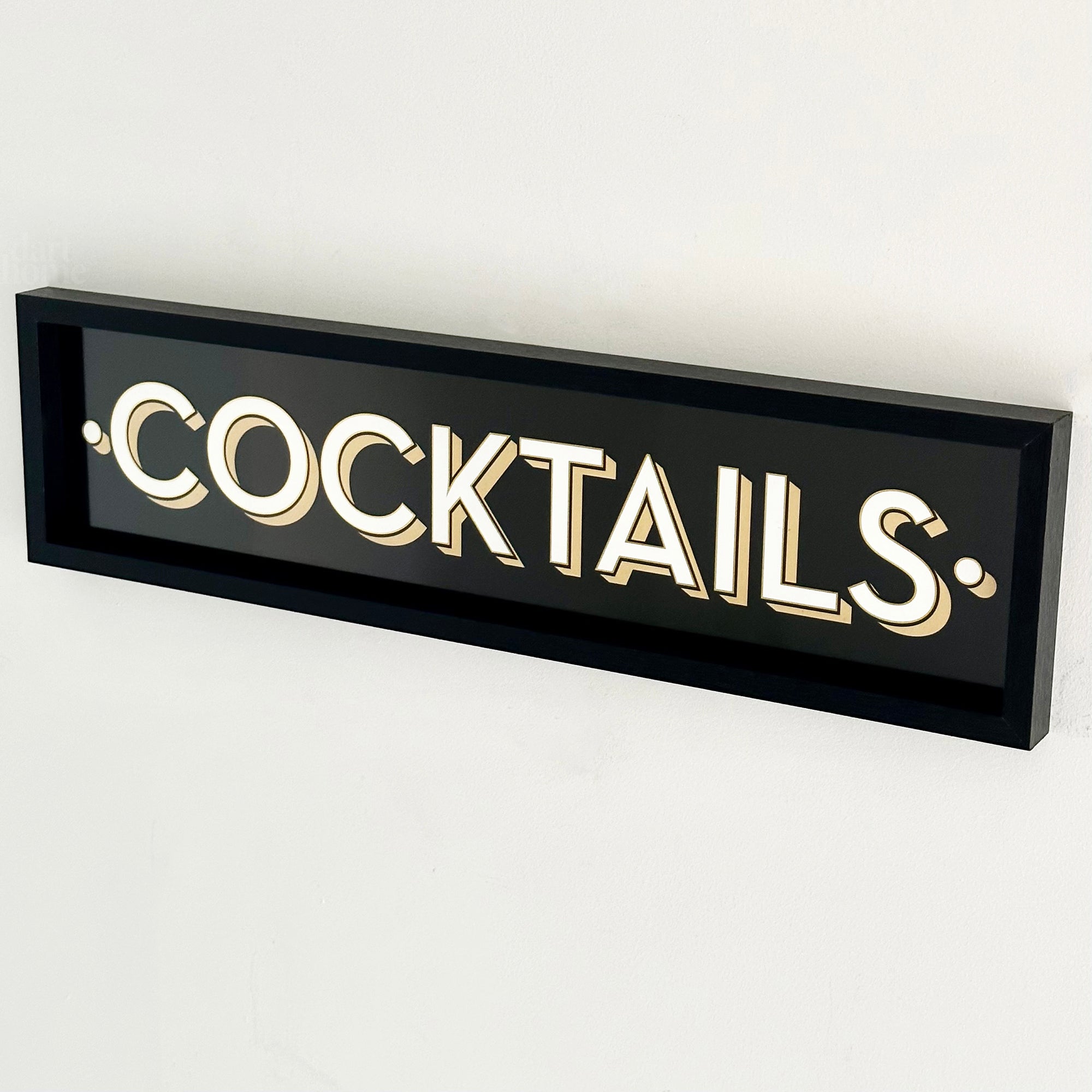 A black framed wall mounted 'COCKTAILS' sign with gold block typography against a white background.