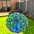 A colorful stained glass suncatcher in the shape of a peacock with blue and green feathers, hanging by a metal chain.