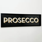 A black framed wall mounted 'Prosecco' bar sign with block typography and a 3D effect, featuring white and gold letters on a black background.